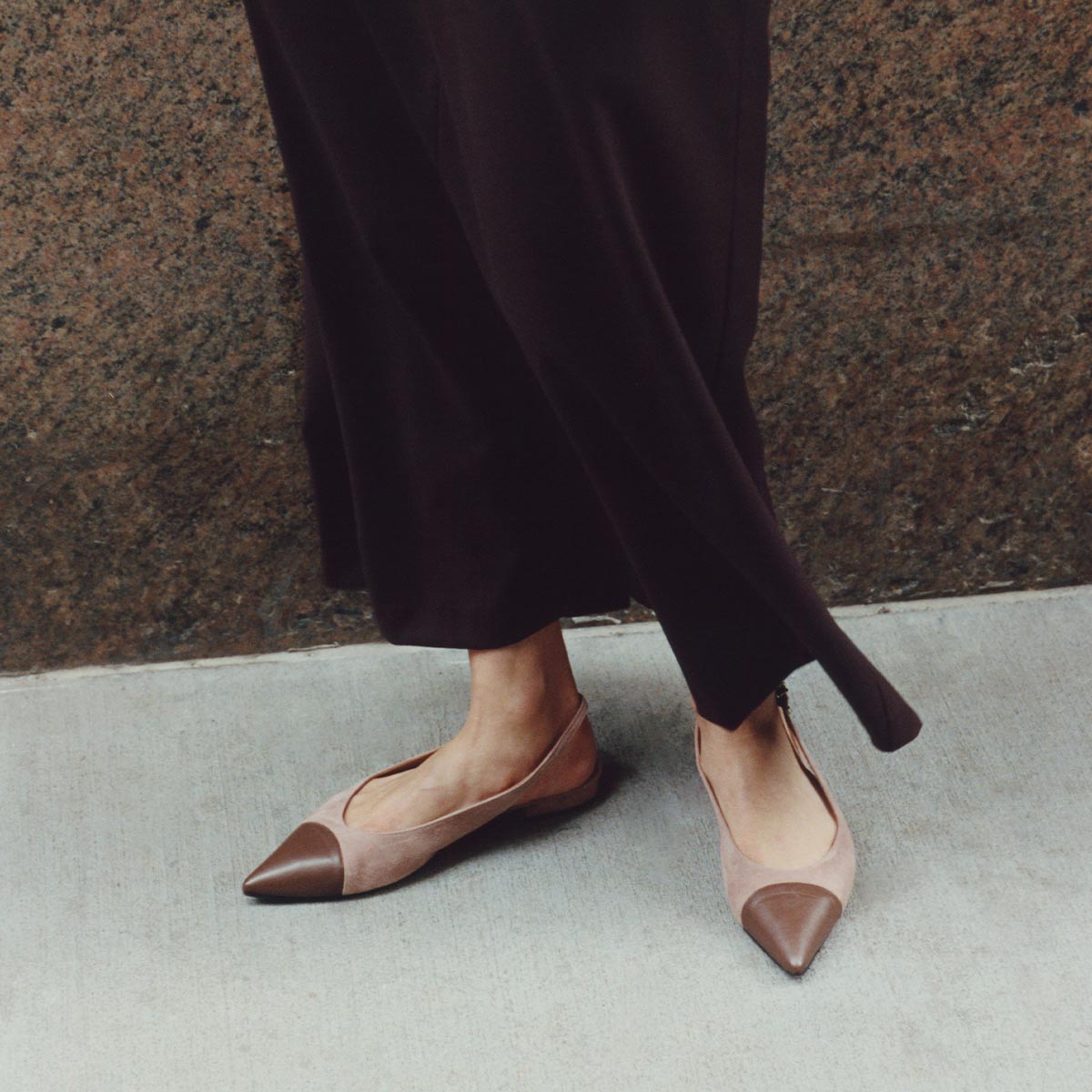 A person wears wide-legged dark pants and The Elise flats in Dusty Rose Suede & Chocolate Nappa by margaux-development, standing on a light concrete sidewalk in front of a granite wall.