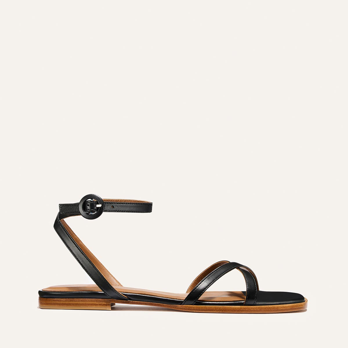 The Margaux Flat Sandal - Black Nappa features a black barely-there design with two thin toe straps, an ankle strap with round buckle, and a brown sole, shown in side profile on a white background for timeless style.