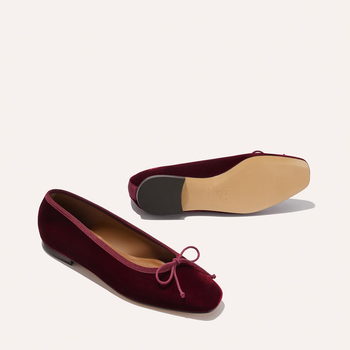 The Fonteyn - Burgundy Velvet by Margaux: handcrafted ballet flats in burgundy velvet with rounded toes, pink bows, and tan leather soles. One comfort-focused shoe stands upright; the other lies on its side to display the sole.