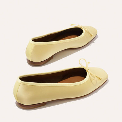 The Fonteyn - Butter Satin by Margaux: handcrafted light yellow ballet flats with rounded toes, small front bows, and brown flat soles, displayed on a plain light background.