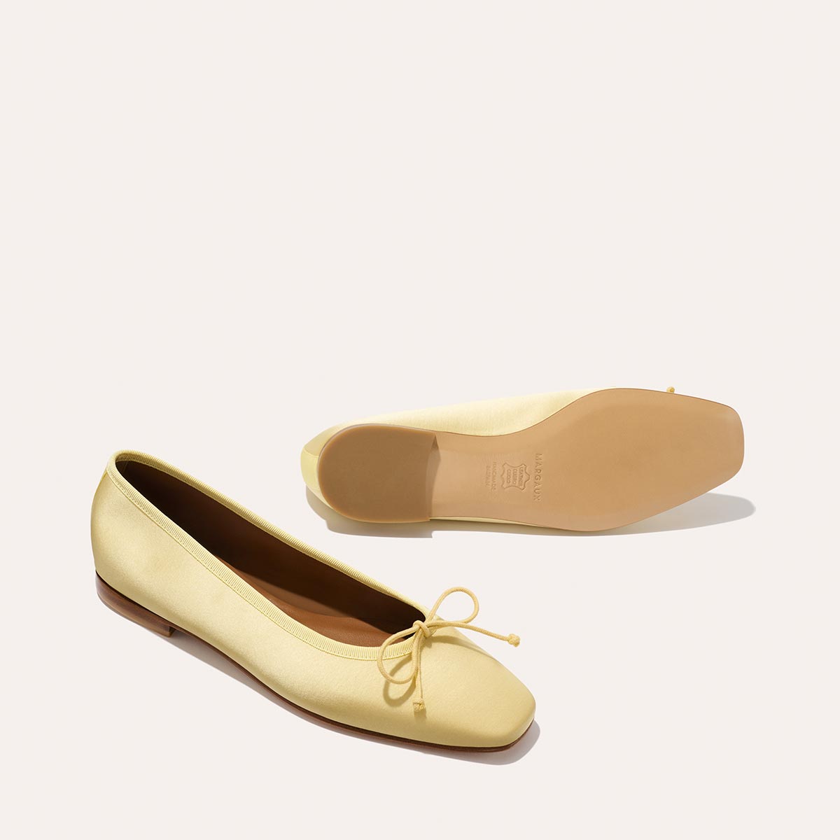 The Fonteyn - Butter Satin by Margaux: a pair of light yellow handcrafted ballet flats with small bows on the toes, shown on a plain light background. One shoe stands upright while the other shows its sole.