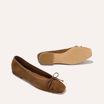 A pair of Margaux's The Fonteyn ballet flats in cedar suede, featuring small front bows. One handcrafted shoe is upright, showing the top and side, while the other rests on its side, displaying the sole, against a plain light background.