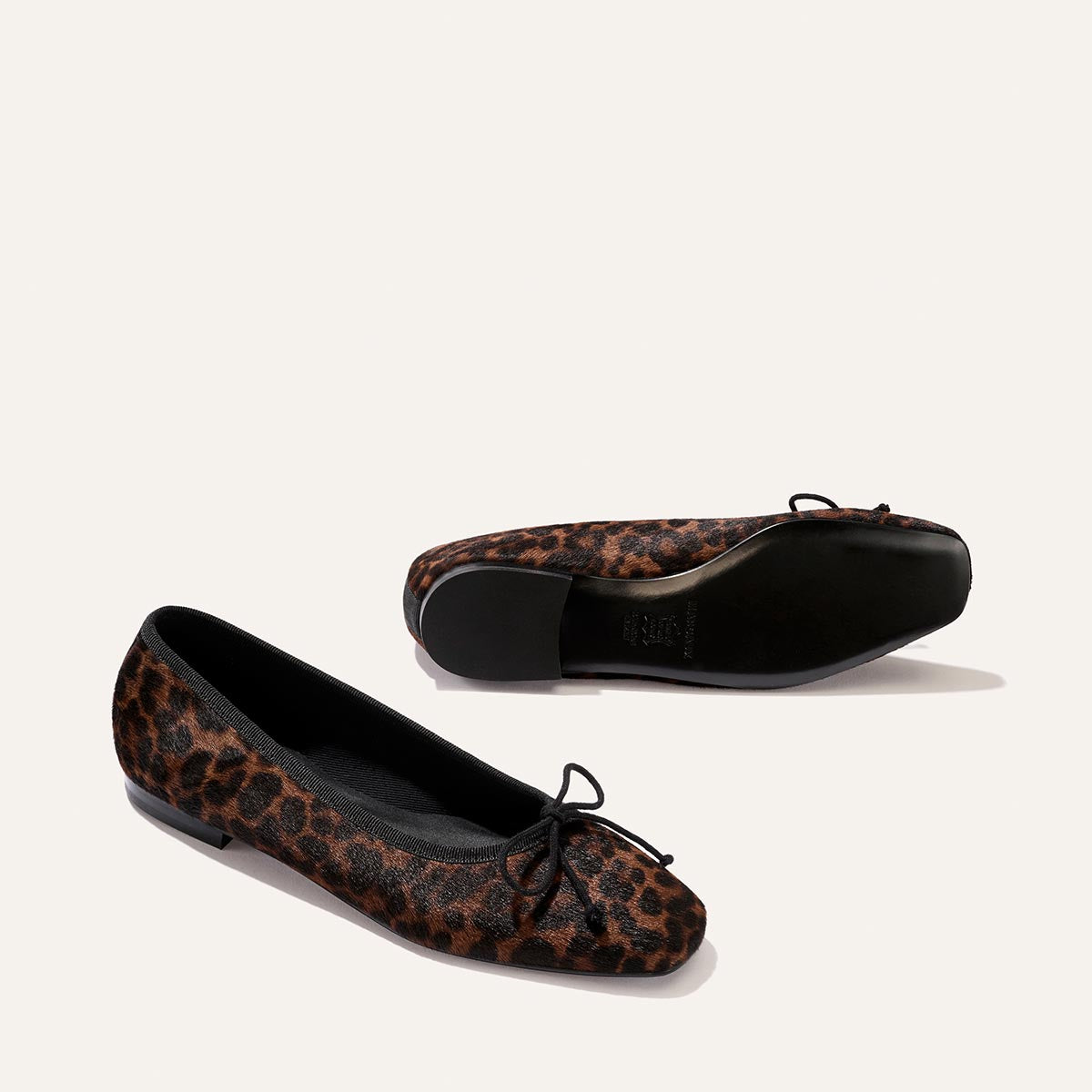 Two pairs of The Fonteyn - Chocolate Leopard Haircalf ballet flats by Margaux, meticulously handcrafted for all-day comfort, showcase black soles and dainty toe bows. Set against a plain, light background, one flat stands upright while the other rests on its side to display the sole.