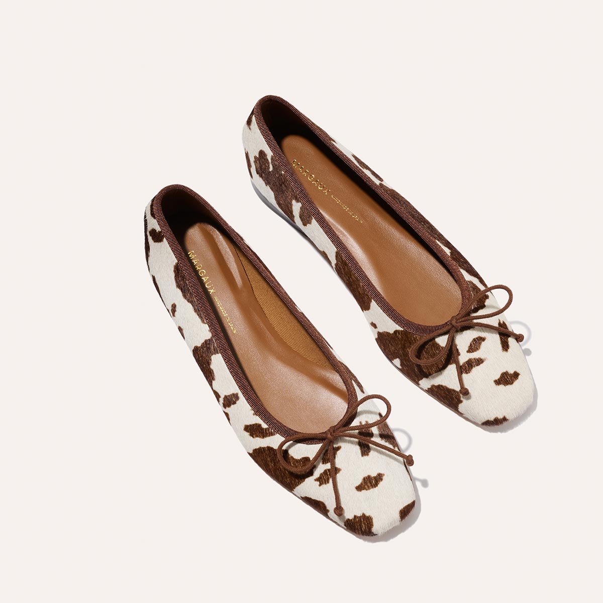The Margaux “The Fonteyn - Cow Print Haircalf” ballet flats feature a chic brown and white cow print with brown bow accents at the toes, shown on a plain white background.