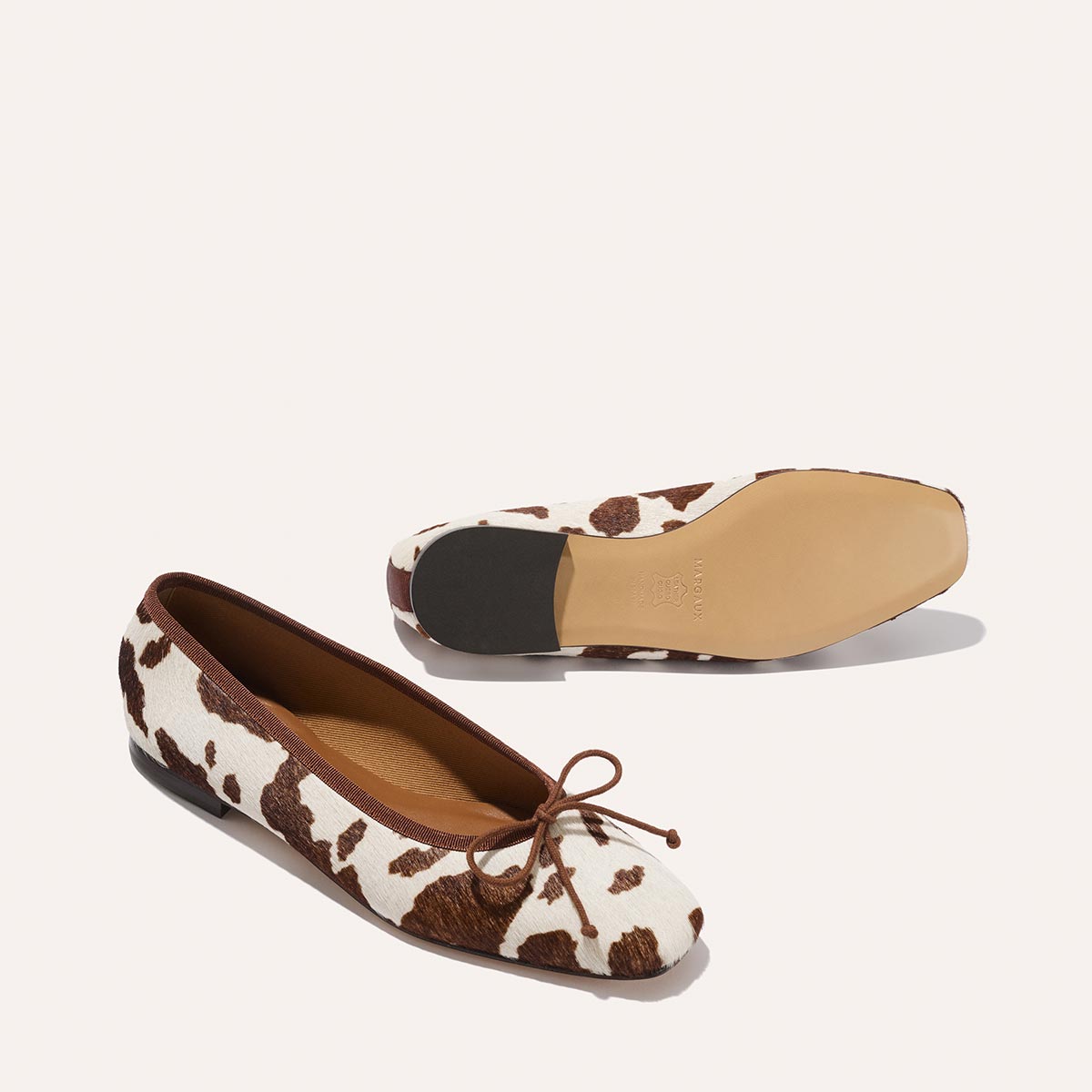 The Fonteyn - Cow Print Haircalf by Margaux: Chic ballet flats in brown and white cow print with brown lining and toe bows. One shoe stands upright, the other rests on its side to show the sole.