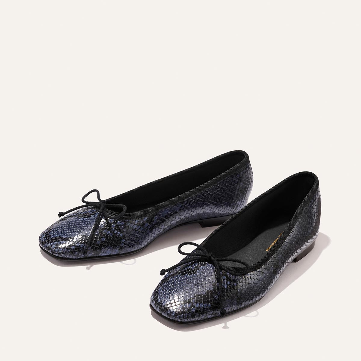 The Fonteyn - Fumo Python Embossed by Margaux: black ballet flats with shiny blue python embossing and black bows on the toes, shown on a light background.