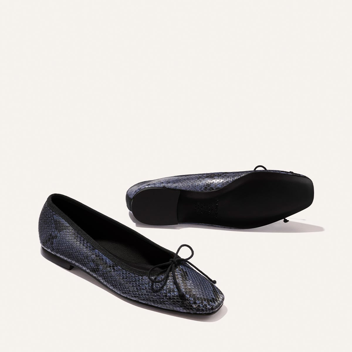 Two Margaux "The Fonteyn - Fumo Python Embossed" ballet flats in black and blue snakeskin pattern with front bows—one upright, one on its side—are displayed on a plain light background.