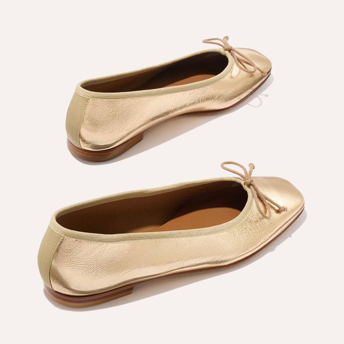 The Fonteyn - Gold Crinkle Nappa by Margaux: a pair of shiny gold ballet flats with small bows on rounded toes and tan soles, displayed on a light background.