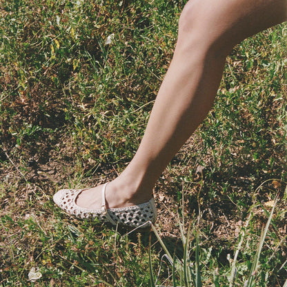 A person wearing Margaux’s The Fonteyn Jane in ecru woven leather, featuring an adjustable strap, steps on grass as sunlight highlights their bare leg and the elegant Mary Jane design.