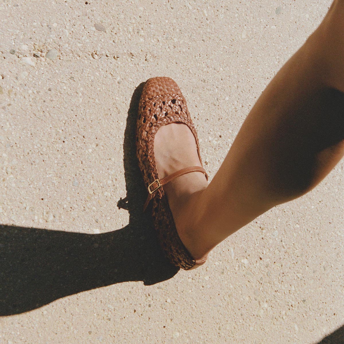 Wearing Margaux's The Fonteyn Jane - Saddle Woven Leather flats with an adjustable strap, someone steps onto a sunlit, textured sidewalk, casting a gentle shadow.