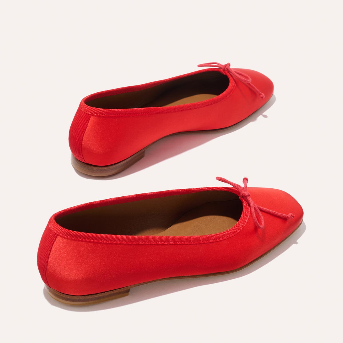 The Fonteyn - Poppy Satin by Margaux are bright red, handcrafted ballet flats with small bows and brown soles, shown on an off-white background for a comfort-focused look.