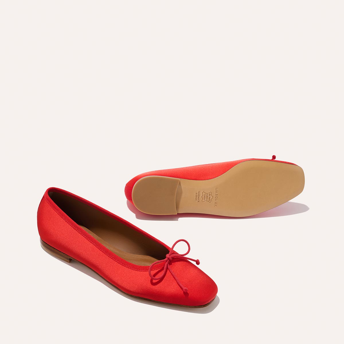 Two Margaux Fonteyn ballet flats in Poppy Satin, each with a small bow—one upright and one flipped to reveal its flat tan sole. The background is plain and light.