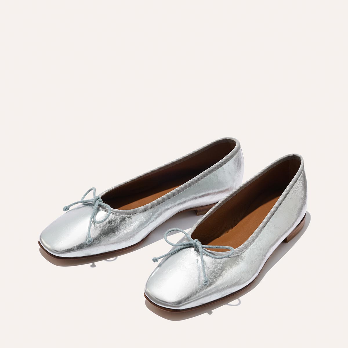 The Fonteyn - Silver Crinkle Nappa by Margaux: shiny silver square-toe ballet flats with small front bows and brown insoles, displayed on a light background.