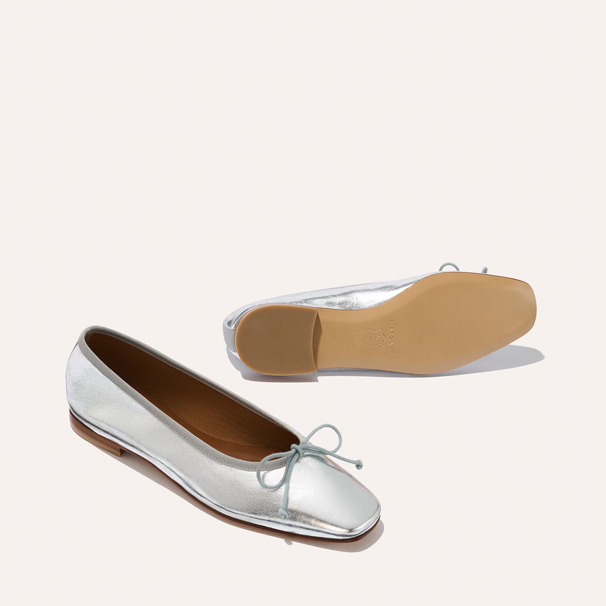 The Fonteyn - Silver Crinkle Nappa ballet flats by Margaux feature shiny silver uppers with a bow on the toe, brown insoles, and tan outsoles. One shoe stands upright while the other rests on its side against a light background.