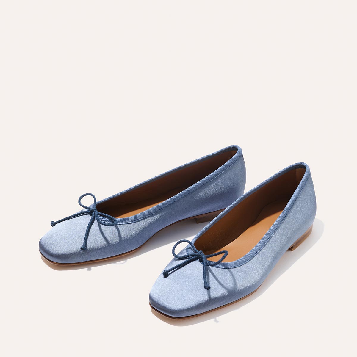 The Fonteyn - Slate Blue Satin ballet flats by Margaux feature square toes, small front bows, and flat tan soles, shown on a light background.