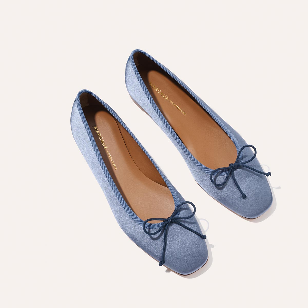 The Fonteyn - Slate Blue Satin by Margaux: sleek ballet flats in light blue satin with delicate toe bows, tan insoles, and a minimalist design, displayed on a light background.