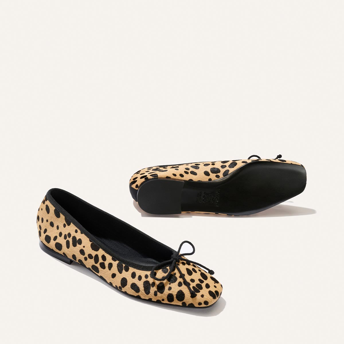 Displayed on a plain white background are two of Margaux's "The Fonteyn - Tan Cheetah Haircalf" ballet flats, featuring a chic cheetah-print design with black soles and delicate black bows at the toes. Crafted with comfort in mind, one flat stands upright while the other lies on its side to showcase the sole.