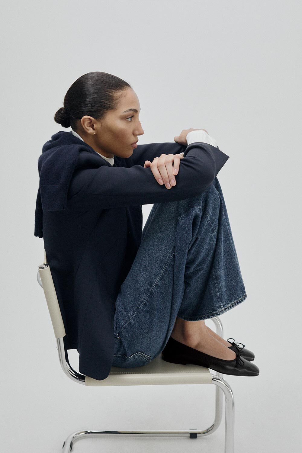 Woman sitting on a chair wearing a dark jacket and jeans against a plain background