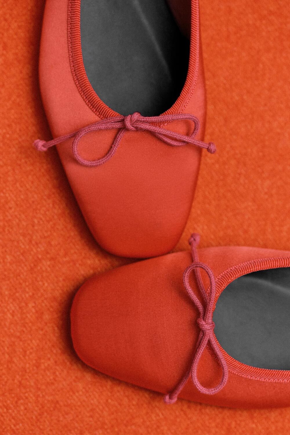 Two red ballet flats with square toes and bow details, placed on an orange textured surface. The shoes are partially overlapping, viewed from above.