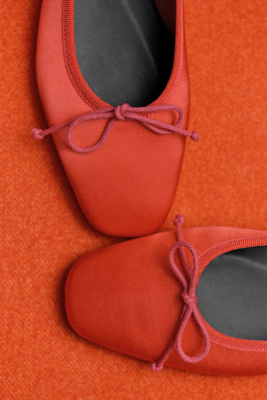 Two red ballet flats with square toes and bow details, placed on an orange textured surface. The shoes are partially overlapping, viewed from above.