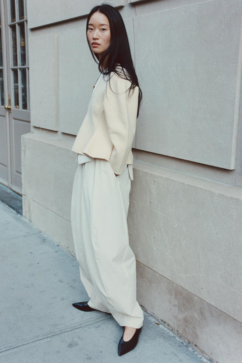 Woman in a beige sweater and white pants standing against a gray wall.