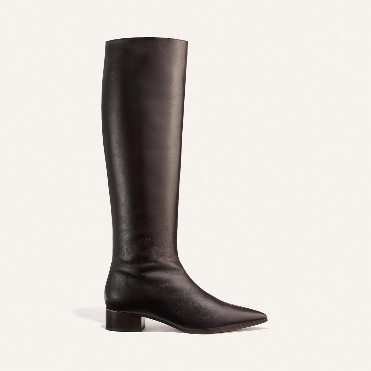The Hayes Knee-High Boot in Dark Chocolate Calf by Margaux is a handmade tall boot crafted from soft leather, featuring a knee-high silhouette with a low block heel, almond toe, and smooth finish. Shown against a light background, it offers a comfortable yet sophisticated look.