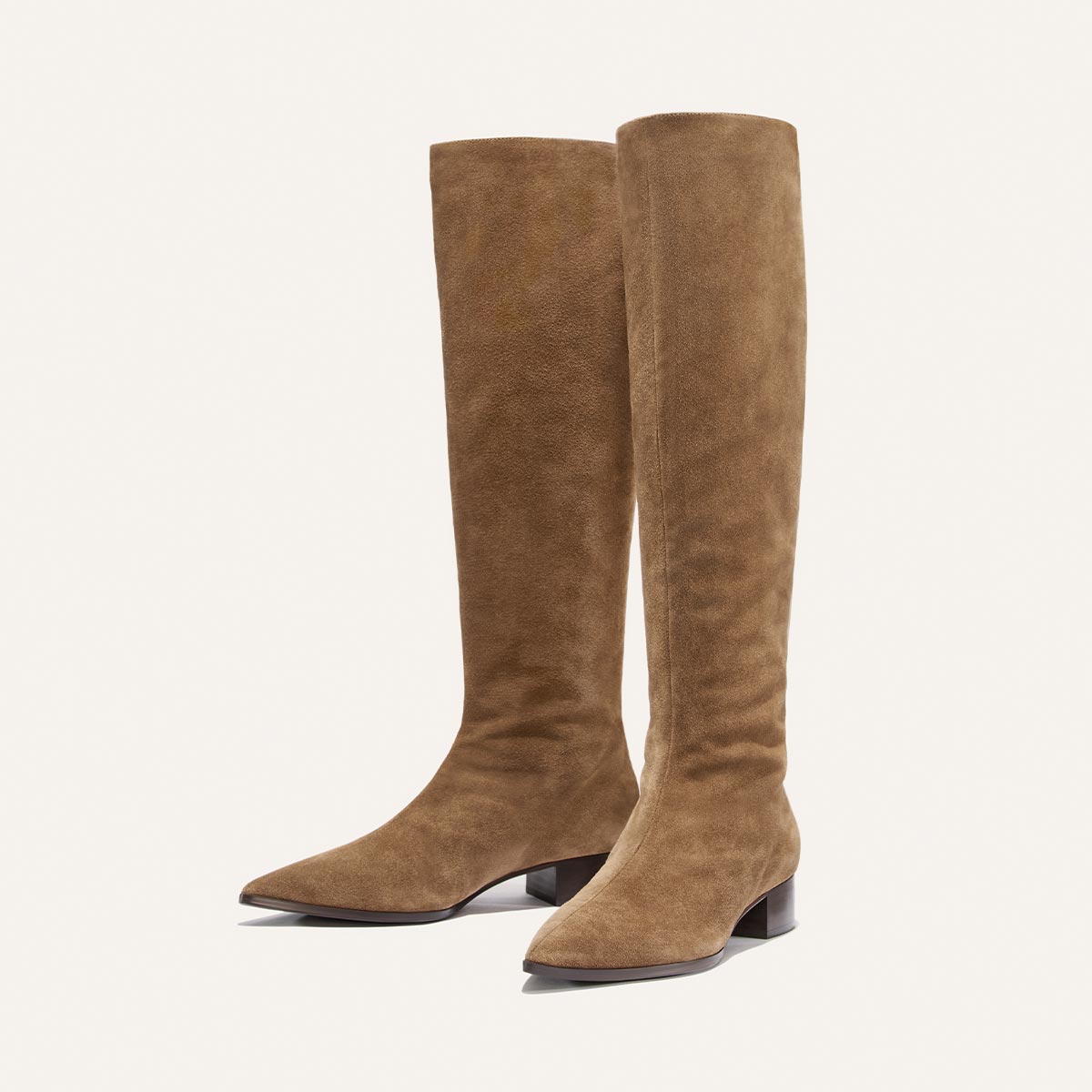 The Hayes Knee-High Boot in Driftwood Suede by Margaux—a pair of knee-high boots crafted from tan suede—features a pointed toe, low block heel, and a flexible sole for effortless movement, elegantly displayed against a plain light background.