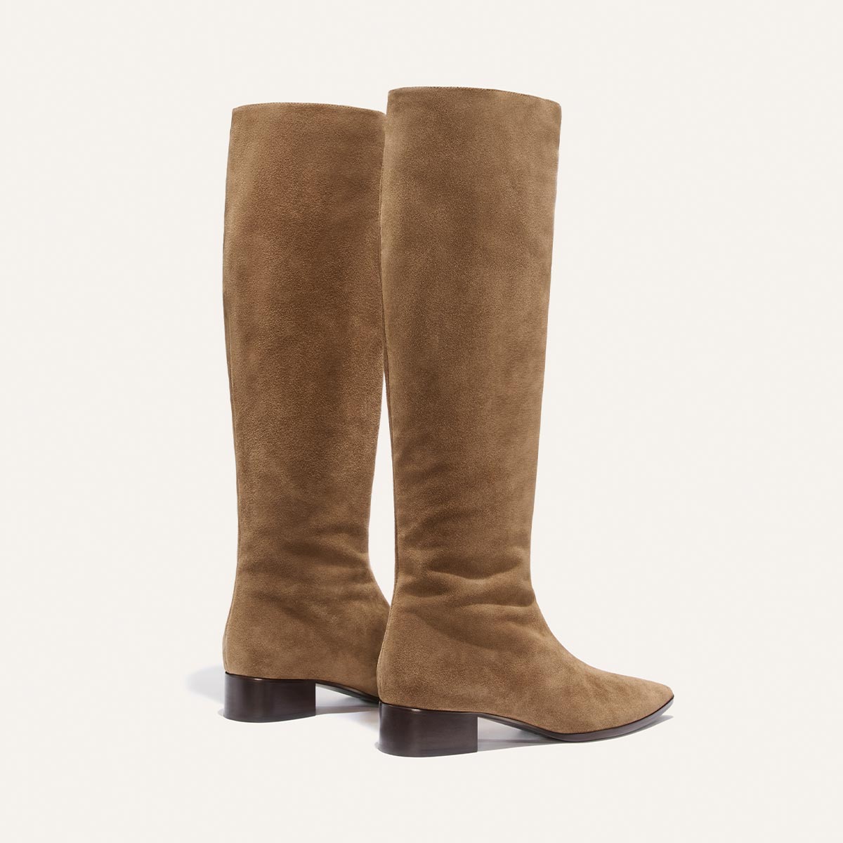 The Hayes Knee-High Boot in Driftwood Suede by Margaux is a pair of tall, tan suede boots with low, dark heels and rounded pointed toes, featuring a flexible sole for all-day comfort, displayed against a plain light background.