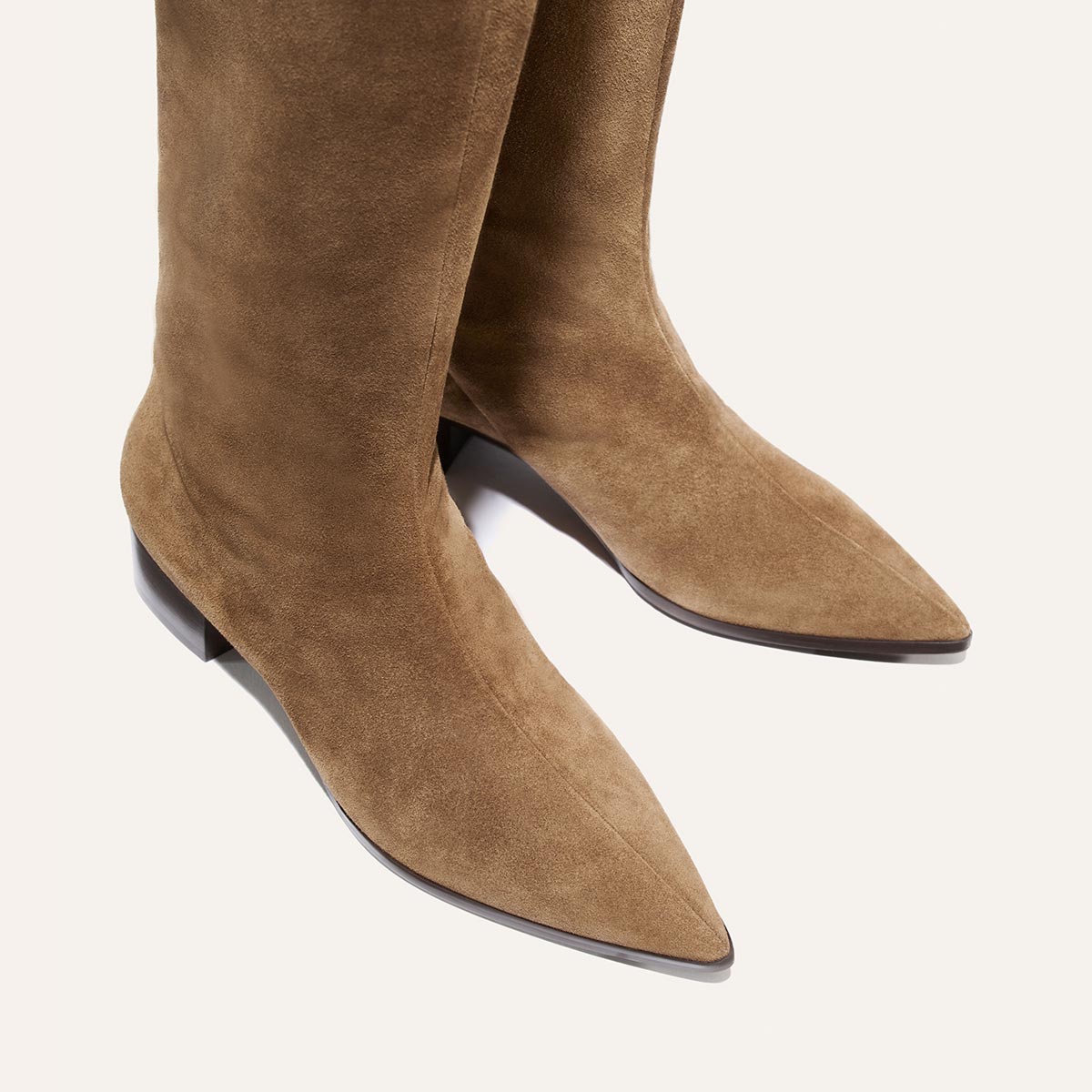 The Hayes Knee-High Boot in Driftwood Suede by Margaux is a pair of tan suede boots with pointed toes and low black heels, featuring a flexible sole for effortless movement, shown against a light background.