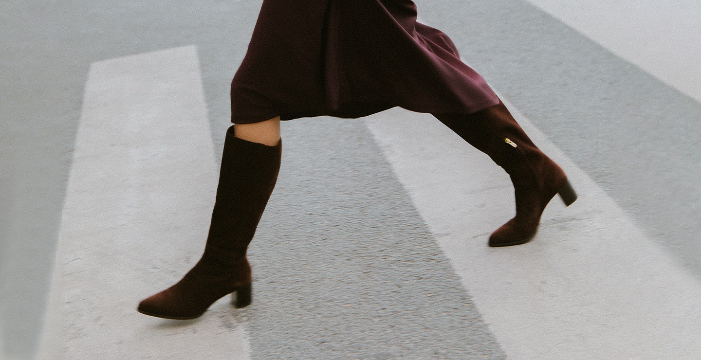 A person wearing a dark purple skirt and matching high-heeled boots is walking briskly across a crosswalk. The photo captures their legs mid-step.