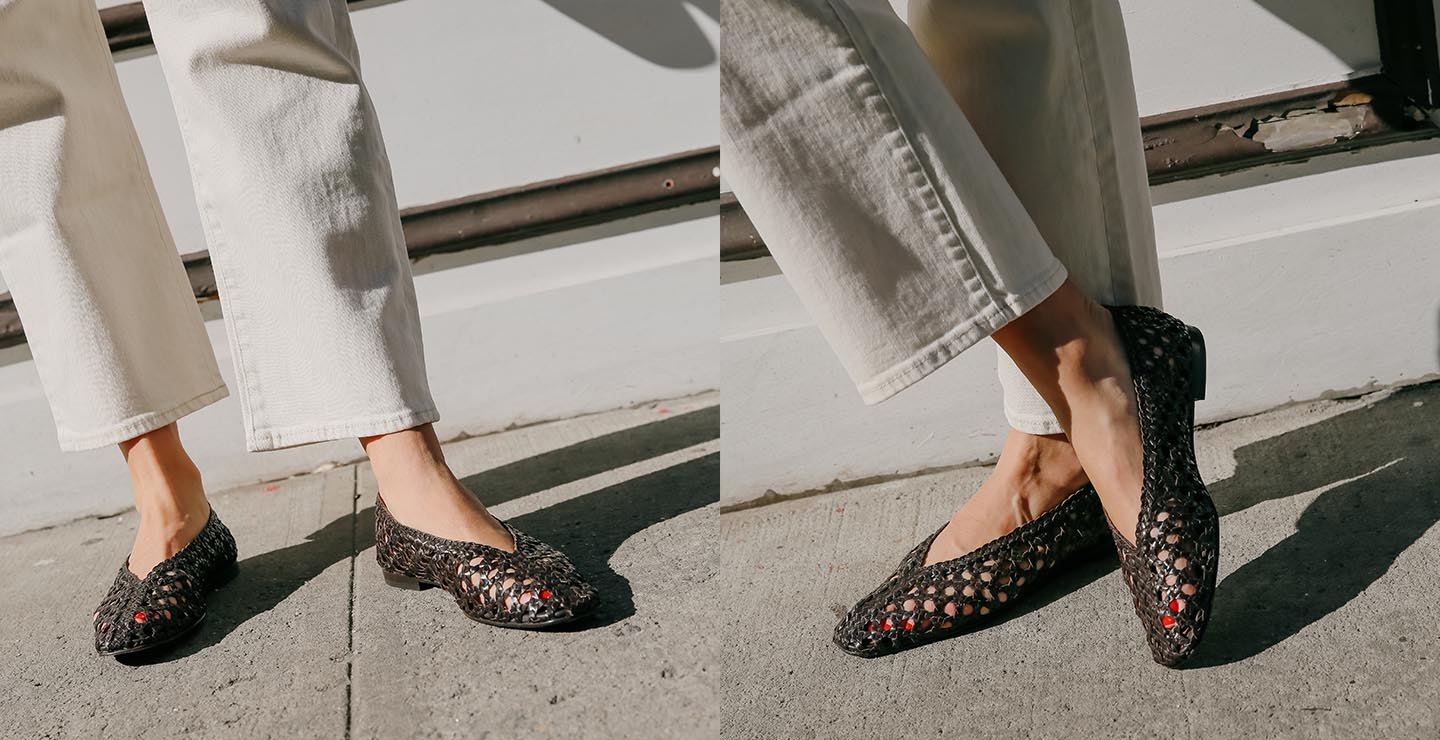 A person wearing cream-colored pants and black woven flats with open toes, standing on a sunny sidewalk. The shoes reveal red-painted toenails.