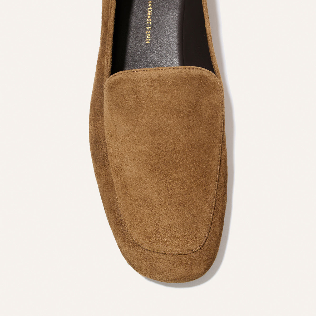 A close-up top view of The James Loafer in Cedar Suede by Margaux showcases its rounded toe, tan suede construction, and visible stitching details against a light background. This handmade loafer is designed with a tailored fit, ensuring exceptional comfort and refined style.