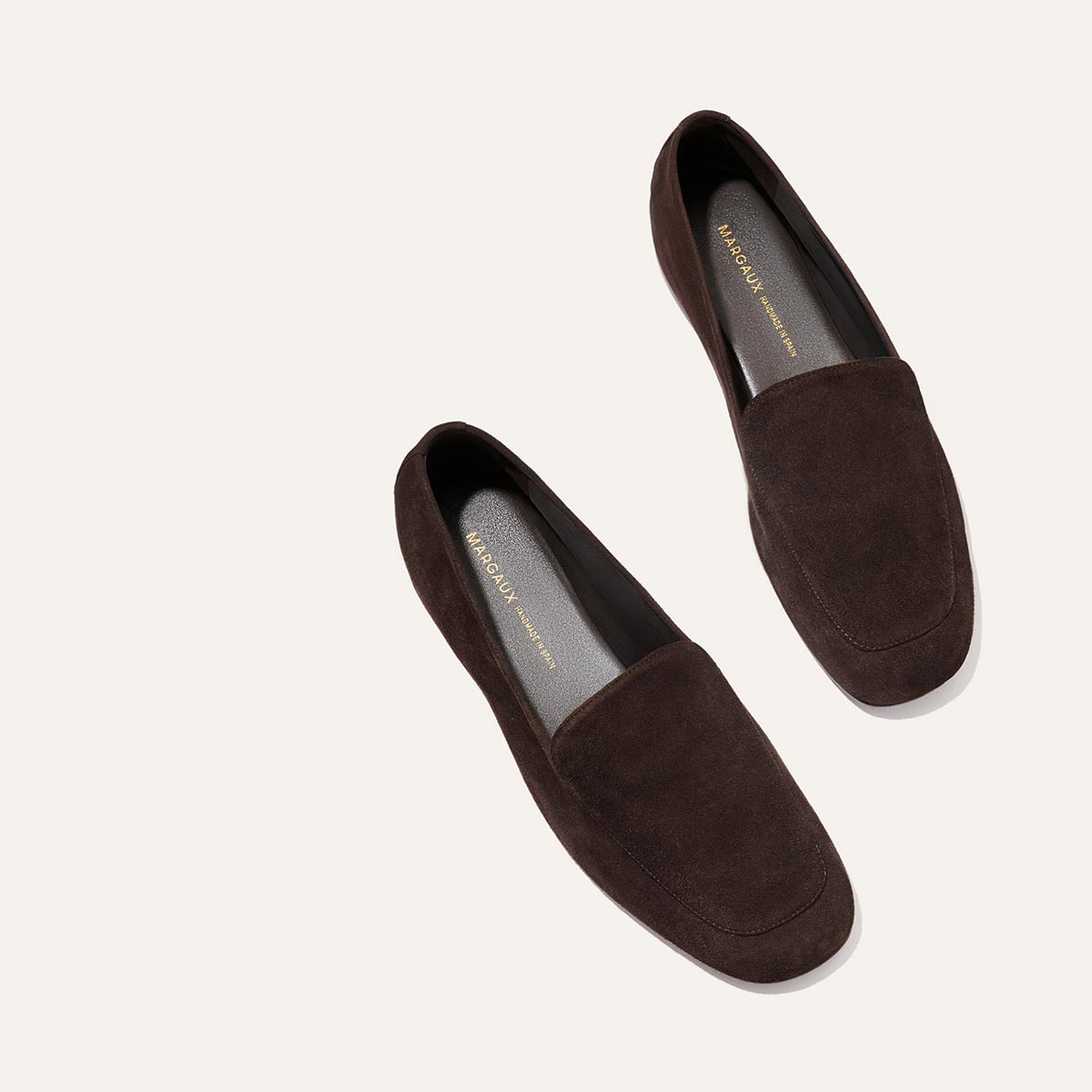 Introducing The James Loafer - Molasses Suede by Margaux: A pair of dark brown suede loafers with a minimalist design, showcased on a light background. Expertly crafted using sacchetto construction, these shoes feature a slightly squared toe and gold Margaux branding on the insoles for an elegant finishing touch.