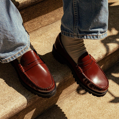 A person stands on outdoor concrete steps in sunlight, wearing light beige socks, blue jeans, and Margaux's The Louisa Loafer First Edition – Hickory Calf with black lug soles and leather-lined insoles.