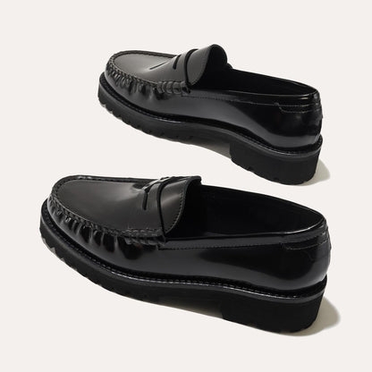 A pair of Margaux’s The Louisa Loafer First Edition in Black Calf, featuring thick lug soles and a penny slot detail, are leather-lined and displayed diagonally on a light background.