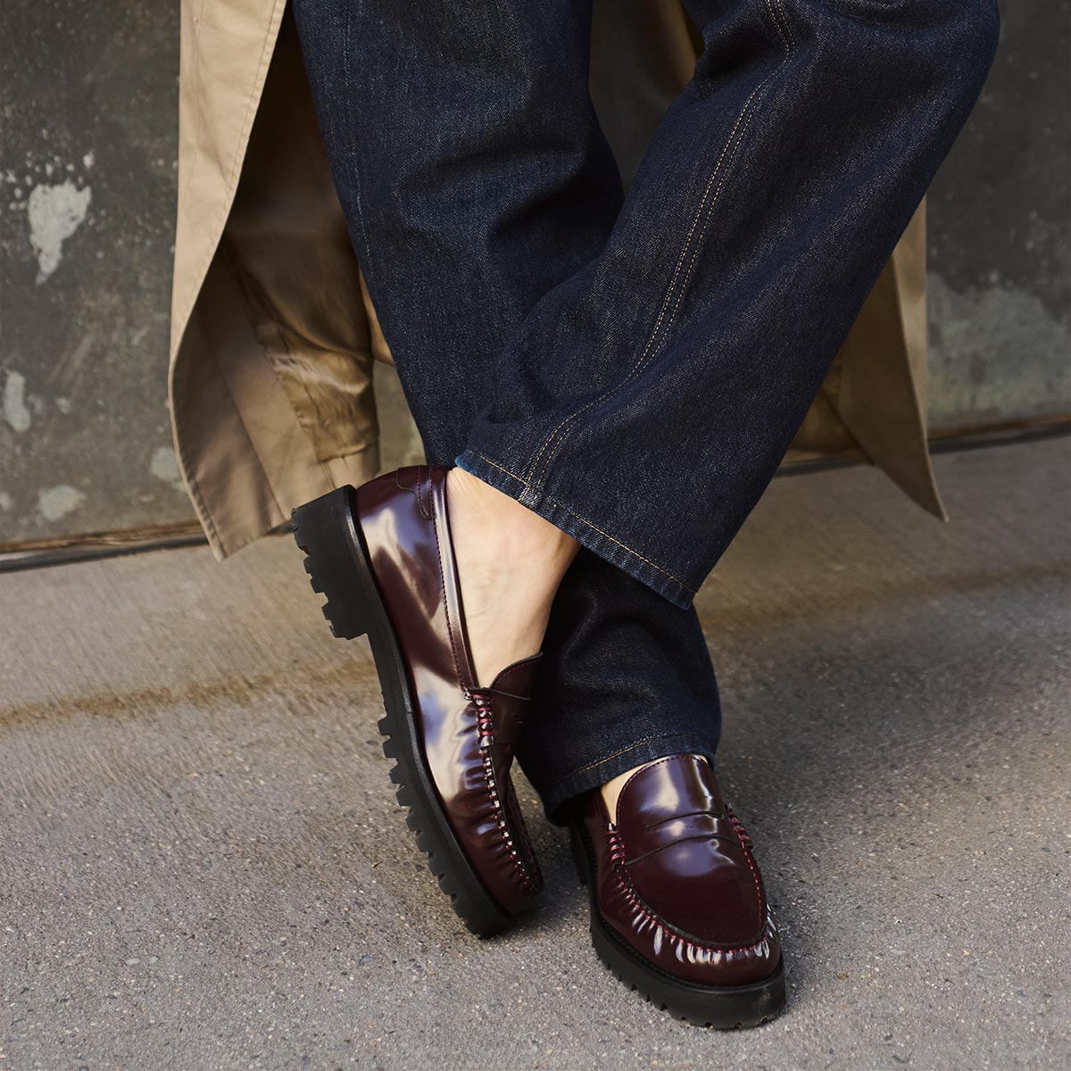 Wearing dark blue jeans, a beige trench coat, and Margaux’s The Louisa Loafer in Burgundy Calf, a person stands on concrete with one foot crossed over the other, highlighting a polished yet comfortable style.