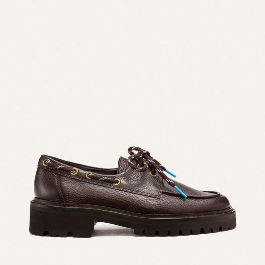 The Marine Loafer - Espresso Tumbled Calf by Margaux is a brown leather loafer featuring a chunky black lug sole, gold eyelets, and black laces tipped with blue aglets, displayed in side profile against a simple backdrop.