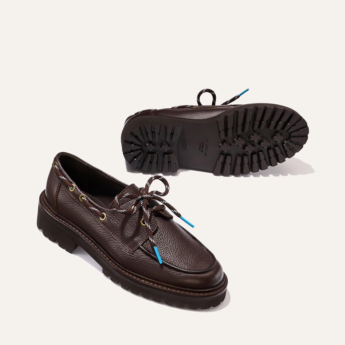 A pair of Margaux’s The Marine Loafer in Espresso Tumbled Calf features brown leather uppers, black rugged soles, yellow eyelets, and patterned laces. One shoe stands upright to highlight the side profile while the other is flipped over to showcase the textured sole.
