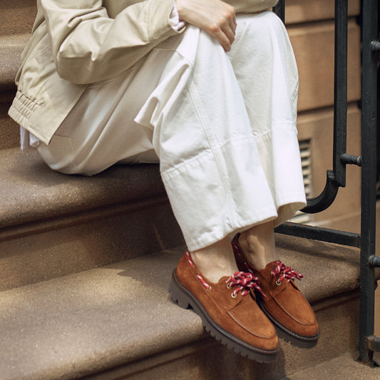 A person sits on outdoor steps wearing off-white pants and Margaux’s The Marine Loafer in Saddle Suede, featuring chunky soles and red patterned laces. Only the lower half of the body is visible.