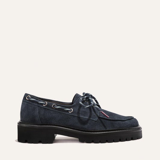 Margaux’s The Marine Loafer in vintage navy suede features silver eyelets, blue-and-white laces, and a chunky black lug sole, shown against a light background.