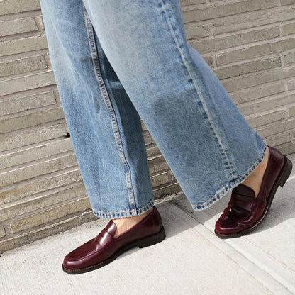 Wearing loose-fitting blue jeans and shiny The Marlowe Loafer - Burgundy Calf by margaux-development, a true closet staple, a person stands on a sidewalk beside a light-colored brick wall.