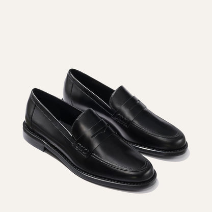 The Marlowe Loafer - Black Calf by margaux-development features classic black leather, rounded toes, low heels, a stitched seam around the upper, and a sleek polished finish. Both shoes are angled outward on a plain light background.