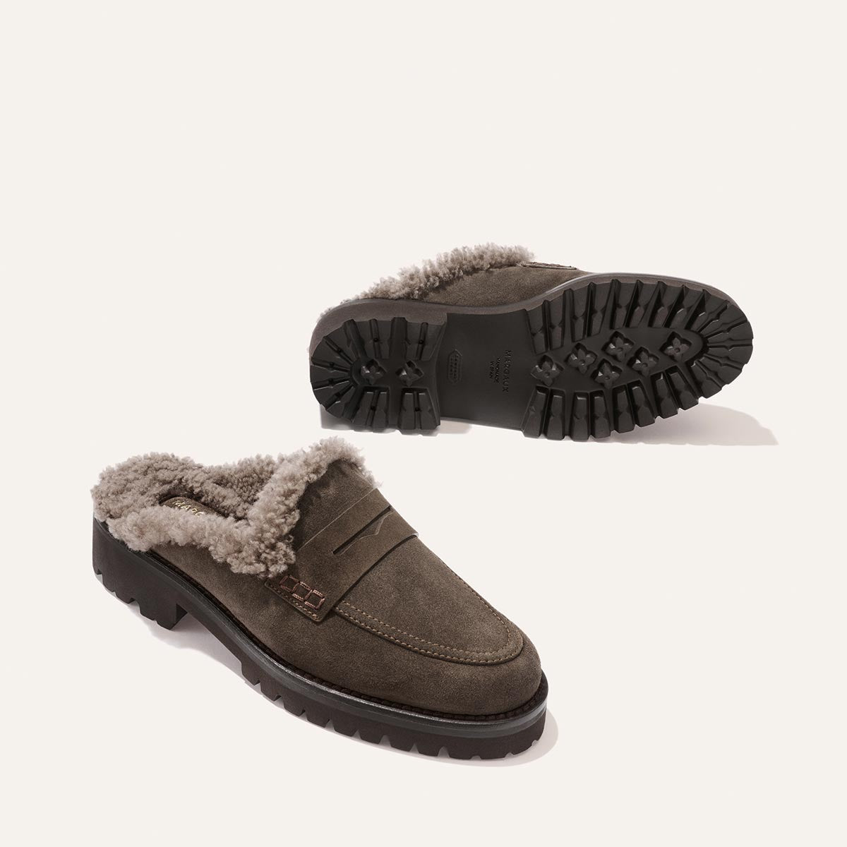 A pair of The Marlowe Mule slip-ons by Margaux, crafted from English brown suede and lined with shearling, featuring chunky black treaded soles, displayed on an off-white background.