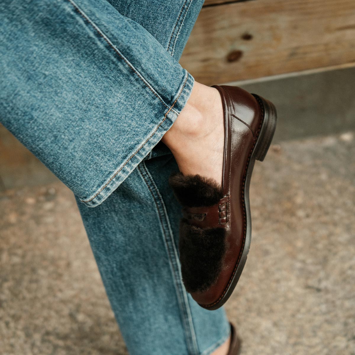 A person in blue jeans and Margaux's The Marlowe Loafer in Mahogany Calf and Shearling sits cross-legged on a stone surface by a wooden bench.