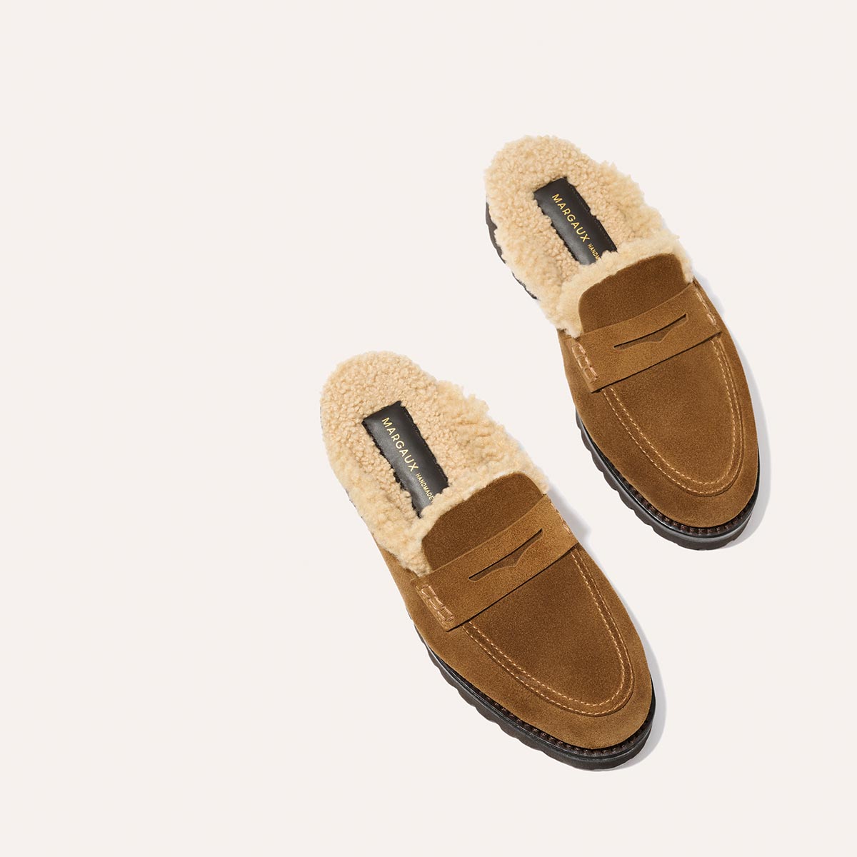 Margaux’s The Marlowe Mule in walnut suede features beige shearling lining, black soles, and visible brand labels on the insoles. The pair is displayed on a light background.
