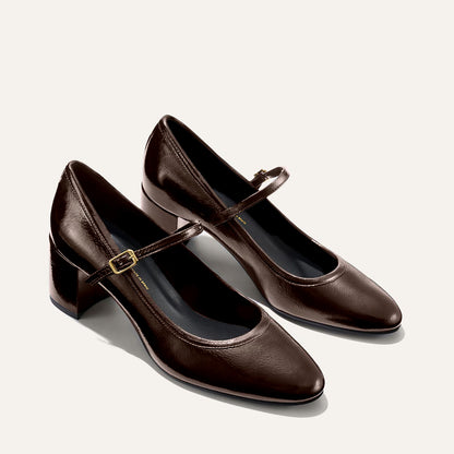 The Mira Jane - Cocoa Crinkle Patent by margaux-development features dark brown gloss Mary Jane shoes with low block heels and thin buckled straps, shown on a light background.