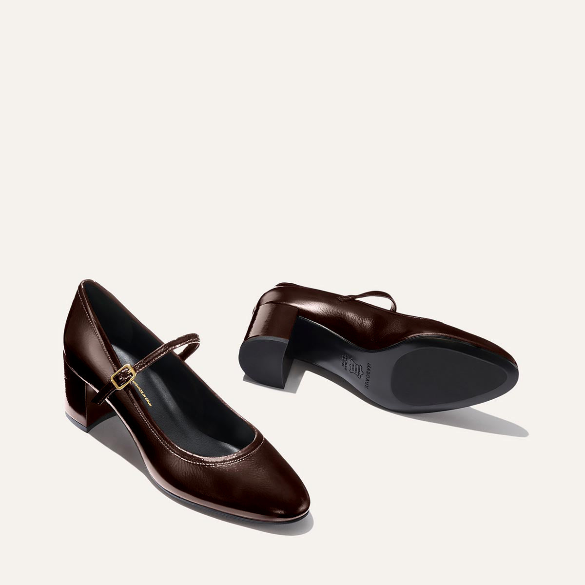 The Mira Jane - Cocoa Crinkle Patent by margaux-development: dark brown gloss Mary Jane shoes with low block heels and thin buckle straps, shown on a light beige background—one upright, one on its side.