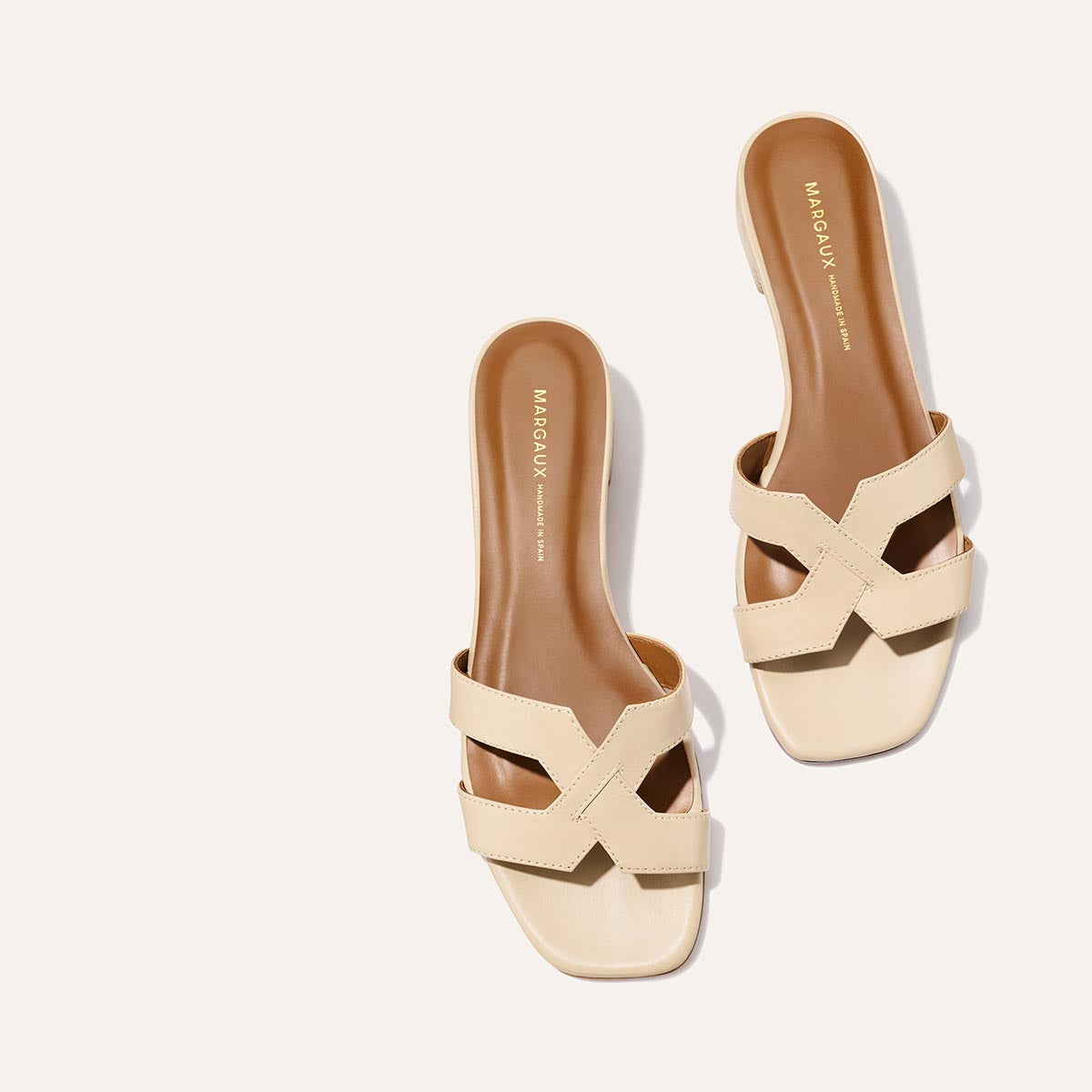 A pair of The MX 35 - Latte Calf sandals by Margaux, featuring beige crisscross straps, brown insoles, and a walkable block heel, displayed on a light background.