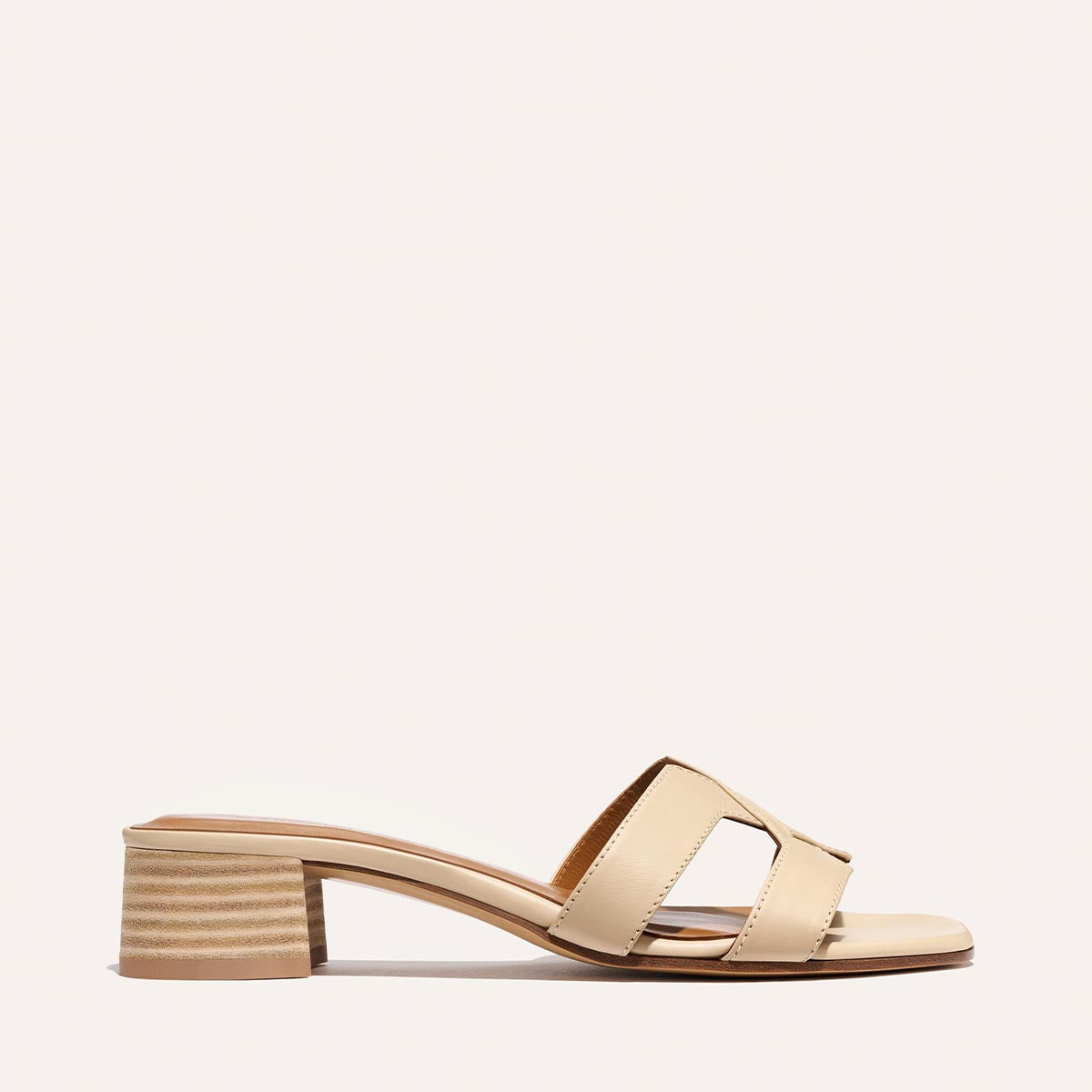 The MX 35 - Latte Calf by Margaux is a beige open-toe sandal with two wide cutout straps and a low block heel. Its minimalist, modern style stands out against a simple, light backdrop.