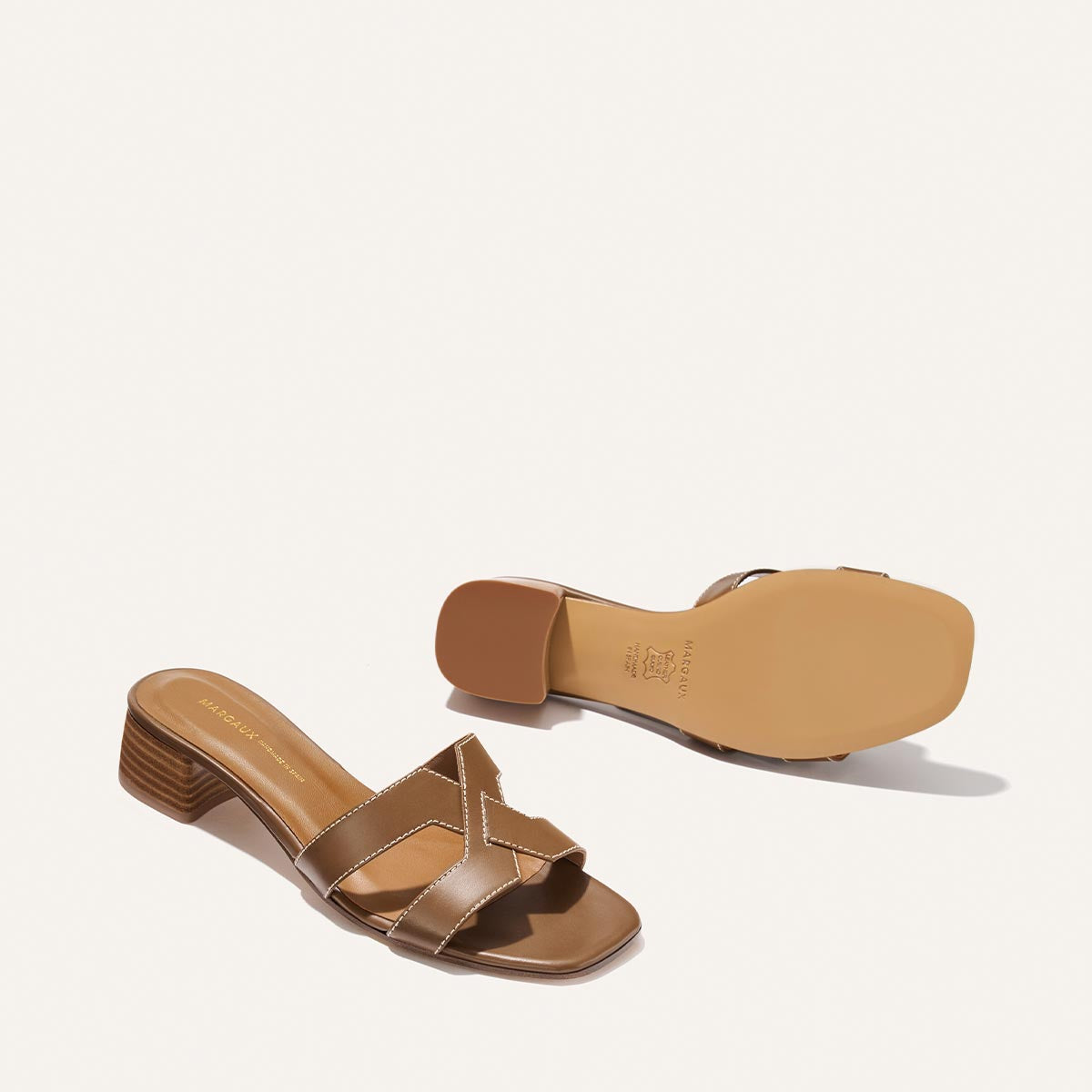 Two MX 35 - Mocha Calf heeled sandals by Margaux are displayed: one upright to show the crisscross straps, one flipped to highlight the sole. Both feature a low block heel and rest against a clean white background.